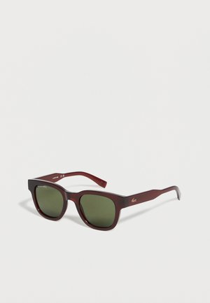 Lunettes de soleil carrées rouge foncé avec verres verts et petit logo argenté sur la branche, sur fond blanc.