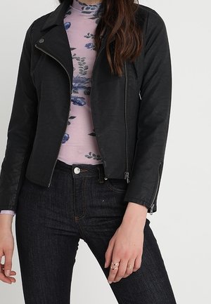 Kobieta w czarnej skórzanej kurtce zapinanej na zamek, na różowym golfie w kwiaty, w parze z ciemnoniebieskimi dżinsami z wysokim stanem i pierścionkami na prawej ręce.