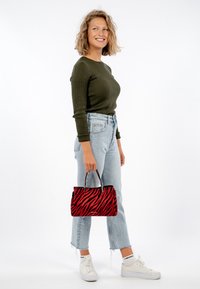 Rød- og sort tigerstribet håndtaske med korte hanke, lædertekstur og sølvhardware, holdt af en person i grøn top og lyse jeans.