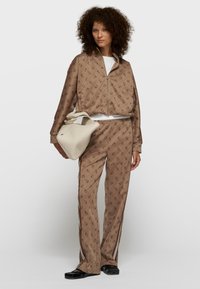 Brązowy dres z powtarzającym się wzorem logo, składający się z kurtki na zamek i spodni o prostych nogawkach. Do kompletu dołączona jest beżowa, fakturowana torba.