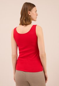 Top rosso senza maniche con una texture liscia e scollatura rotonda. Indossato con pantaloni beige chiaro. Design semplice senza motivi o dettagli.