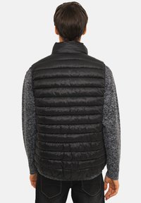 Veste sans manches noire matelassée avec texture matelassée, col montant et design léger. Portée par-dessus un pull gris en maille, accompagnée d'un jean foncé.