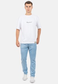 Witte T-shirt met korte mouwen en zwart scriptlogo in het midden, gecombineerd met lichtblauwe, gestructureerde spijkerbroek. Model draagt witte sneakers met rode accenten.