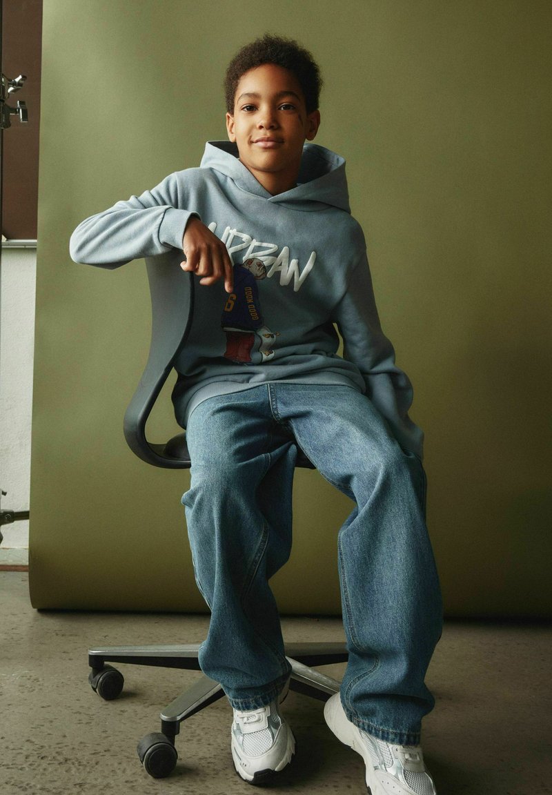 Garçon portant un sweat à capuche bleu, un jean et des baskets blanches assis sur une chaise de bureau noire devant un fond vert uni.