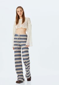 Gehaakte witte crop top met opengebreid patroon; wijde broek met zigzagprint in bruine, beige en blauwe tinten; platte schoenen.