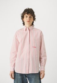 Camicia bianca con strisce verticali rosse, maniche lunghe, colletto con bottoni e ricamo del logo rosso sul petto. Indossata con jeans in denim blu.
