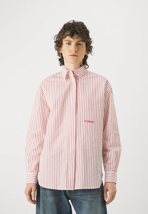 Camicia bianca con strisce verticali rosse, maniche lunghe, colletto con bottoni e ricamo del logo rosso sul petto. Indossata con jeans in denim blu.