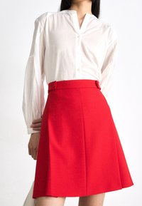 Jupe évasée rouge avec double rangée de boutons sur les côtés, associée à une chemise blanche à boutons, col cranté et manches froncées.