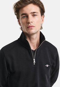Black zip-up pullover made of soft fabric. Features a high collar, silver zipper, and small embroidered logo on the chest.