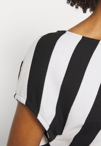 Rechte Schulter einer Person, die ein schwarz-weiß gestreiftes kurzärmliges Oberteil trägt, mit lockigem Haar, das am Nacken sichtbar ist.