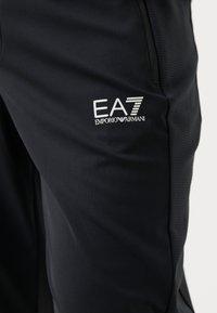 Fekete sportos nadrág sima, rugalmas anyagból, bal combján egy kisméretű fehér EA7 Emporio Armani logóval.
