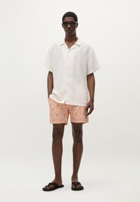 Chemise blanche à manches courtes avec des boutons, shorts orange à motifs affichant un design géométrique, et tongs noires. Esthétique épurée et minimaliste.