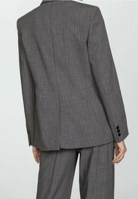 Giacca da abito grigia a righe con vestibilità sartoriale, spacchi posteriori, bottoni neri ai polsini e texture liscia. Indossata con pantaloni abbinati.