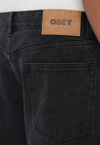 Crne traperice od denima s kožnom etiketom s utisnutim "OBEY". Izrađene su s stražnjim džepovima i standardnim šavovima.