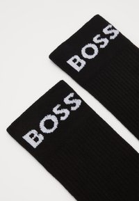 Calcetines negros con textura acanalada y logotipo blanco "BOSS" en la parte superior. El material parece estar tejido y es suave. Diseño sencillo con un aspecto clásico.