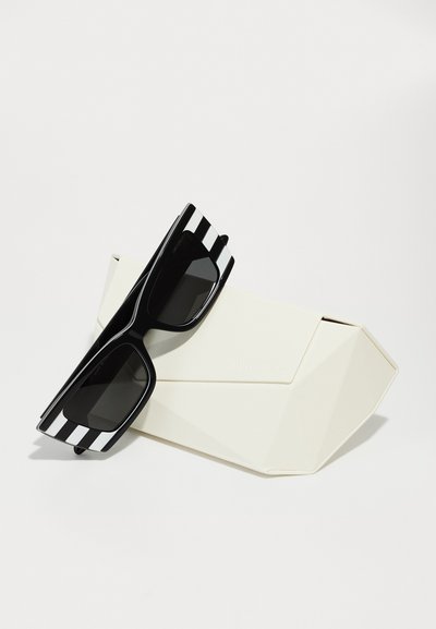 Black sunglasses with white stripes on the arms, placed on a geometric white case. The lenses are dark and rectangular in shape.