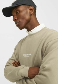 Sudadera color caqui con un cuello redondo, con la impresión "Originals Studio" en blanco. Lleva una camisa blanca debajo. Gorra oscura usada al revés.