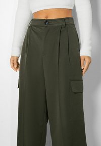 Breda cargo-byxor i olivgrönt, med knappstängning fram, veck och sidofickor för funktionalitet. Slät textur.
