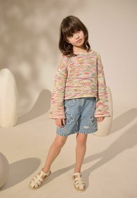Pull en tricot avec des motifs roses, jaunes et verts ; short en denim bleu avec broderie florale ; sandales blanches à lanières.