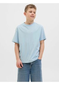 Světle modré bavlněné tričko s krátkými rukávy a kulatým výstřihem, s bílým nápisem "Originals Studio" na prsou. Nosené s modrými džíny.