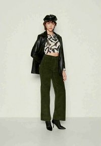 Femme portant une veste en cuir noir, un crop top imprimé, un pantalon taille haute vert olive, des bottes à talons noires et une casquette en cuir noir, se tenant devant un fond pâle.