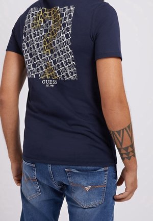 Homme portant un t-shirt Guess bleu marine avec un logo blanc et jaune en forme de grille au dos et un jean bleu, montrant un tatouage sur l'avant-bras gauche.