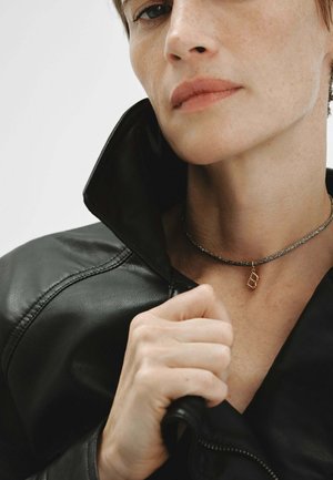 Primer plano de una persona que lleva una chaqueta de cuero negra y un collar con un colgante de letra B en oro, mano sujetando el cuello de la chaqueta.