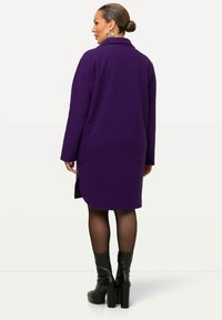 Lång lila rock med krage, sidofickor och rak skärning. Har långa ärmar och slitsar i botten. Bärs med svarta platåstövlar.