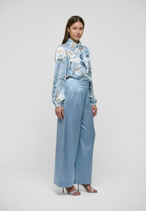 Vrouw draagt een lichtblauw bloesje met bloemenpatroon en striksluiting bij de hals, gecombineerd met wijde satijnen broekspijpen en zilveren sandalen met hoge hak.