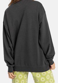 Sudadera de gran tamaño en gris oscuro, con cuello redondo, mangas largas y puños acanalados. Textura notablemente suave, combinada con pantalones verdes de patrón.