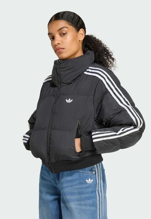A young woman poses wearing a black puffer jacket with white stripes, paired with blue jeans featuring similar stripes, showcasing a sporty style.