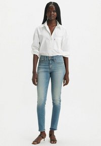 Camisa blanca de botones sobre unos jeans ajustados de un azul claro, con una suave textura de denim, un ligero desgaste y sandalias negras de tiras.