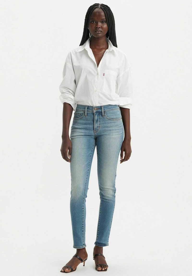 Camisa blanca de botones sobre unos jeans ajustados de un azul claro, con una suave textura de denim, un ligero desgaste y sandalias negras de tiras.