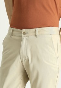 Pantalones beige con una textura suave, que cuentan con un cierre de botón, presillas para cinturón y bolsillos frontales. La tela parece ligera y casual.