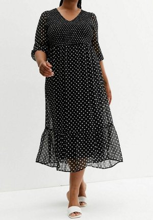 Frau trägt ein schwarzes Midikleid mit weißen Punkten und transparenten Ärmeln, kombiniert mit weißen offenen Slide-Sandalen.