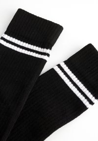 Calze nere fino al ginocchio con due strisce orizzontali bianche nella parte superiore. Il materiale ha una trama a coste. Disposte in posizione distesa su uno sfondo bianco.