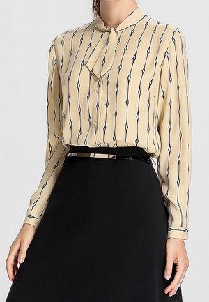 Femme portant un chemisier beige à manches longues avec des rayures en losange noires et une jupe noire avec une fine ceinture à la taille.
