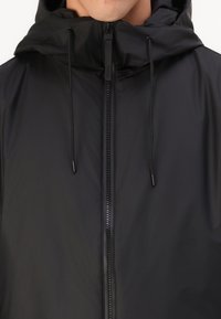 Schwarze wasserdichte Jacke mit Kapuze, ausgestattet mit einem durchgehenden Reißverschluss und verstellbaren Kordeln. Glatte Textur, minimalistisches Design.