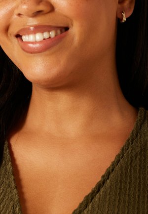 Close-up van het onderste deel van het gezicht, de nek en schouder van een lachend persoon, gekleed in een textuur-olijfgroene top en een kleine gouden oorring.