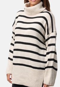 Gestreepte coltrui in crème met zwarte horizontale strepen, voorzien van een losse pasvorm en geribbelde zoom en manchetten. Gemaakt van zacht gebreid materiaal.