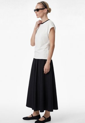 Donna con occhiali da sole neri, top bianco senza maniche, lunga gonna nera, ballerine nere e fascia nera, ritratta di profilo.