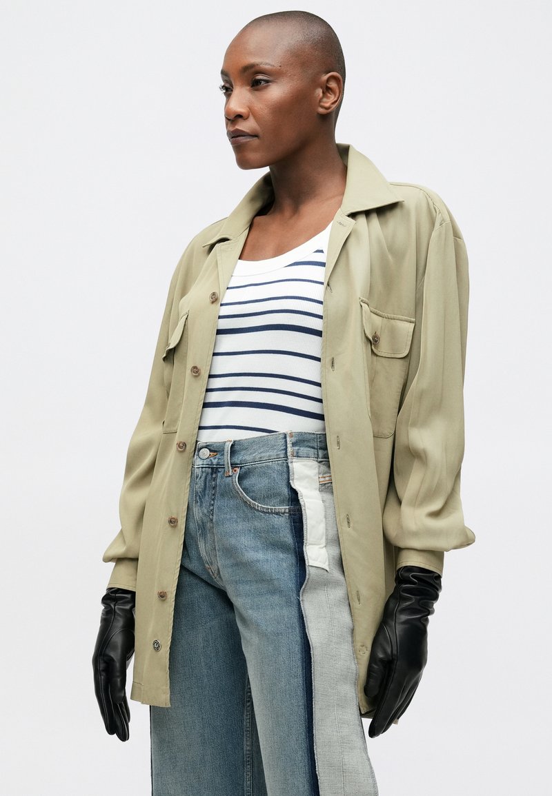 Olivgrön knäppjacka med två framfickor, bärs över en vit och marinrandig topp, tillsammans med blå jeans med sidopaneler och svarta handskar.