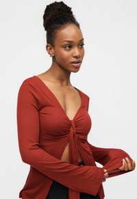 Τοπ μακρυμάνικο σε χρώμα σκουριάς με βαθύ V λαιμόκοψη και κόμπο στο μπροστινό μέρος. Διαθέτει φαρδιά μανίκια και εφαρμοστή γραμμή, κατασκευασμένο από μαλακό, υφασμάτινο ύφασμα με υφή.