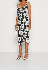 Žena v černých midi šatech s bílým květinovým vzorem, na ramínkách, s předním rozparkem, v bílých vysokých sandálech na podpatku a s malou bílou kabelkou.