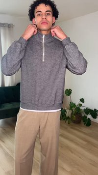 Homme ajustant le col d'un pull gris à motif avec une fermeture éclair jusqu'au quart, debout dans une pièce avec un sol en bois clair, un canapé vert et des plantes en pot.