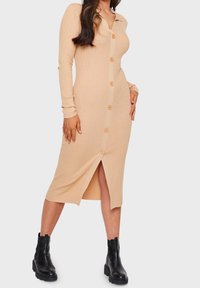 Robe midi en tricot côtelé beige avec boutonnage sur le devant, manches longues et fente latérale ; associée à des bottes noires à talons épais.