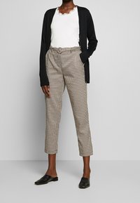 Cardigan noir, haut blanc avec encolure à volants, pantalon à carreaux marron, et mocassins noirs. Le pantalon est ceinturé avec une boucle ronde.