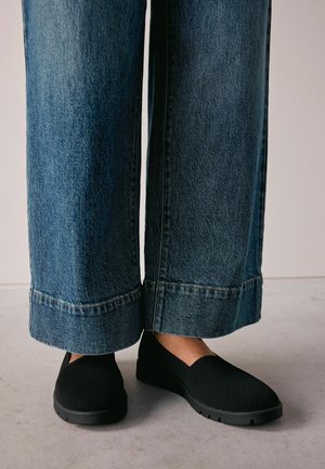 Black slip-on shoes with a textured mesh upper, paired with wide-leg denim jeans featuring a raw hem and contrasting stitching.