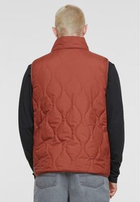 Red quilted vest with high collar and wavy stitching pattern. Worn over a black long-sleeve shirt. Gray jeans are partially visible.