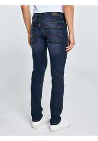 Bărbat purtând blugi slim-fit de culoare albastru închis și adidași albi, stând cu spatele către înainte pe un fundal simplu, deschis la culoare.